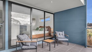 A modern bedroom with a glass door leading to a balcony, a bed with pillows, and two chairs. - Saltwater Apartments - 2 Bedroom (Eden)