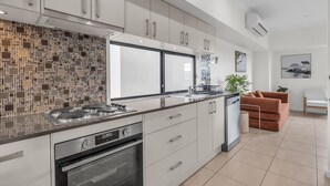 A modern kitchen with a tiled backsplash, stainless steel appliances, and a large window. - Saltwater Apartments - 2 Bedroom (Eden)