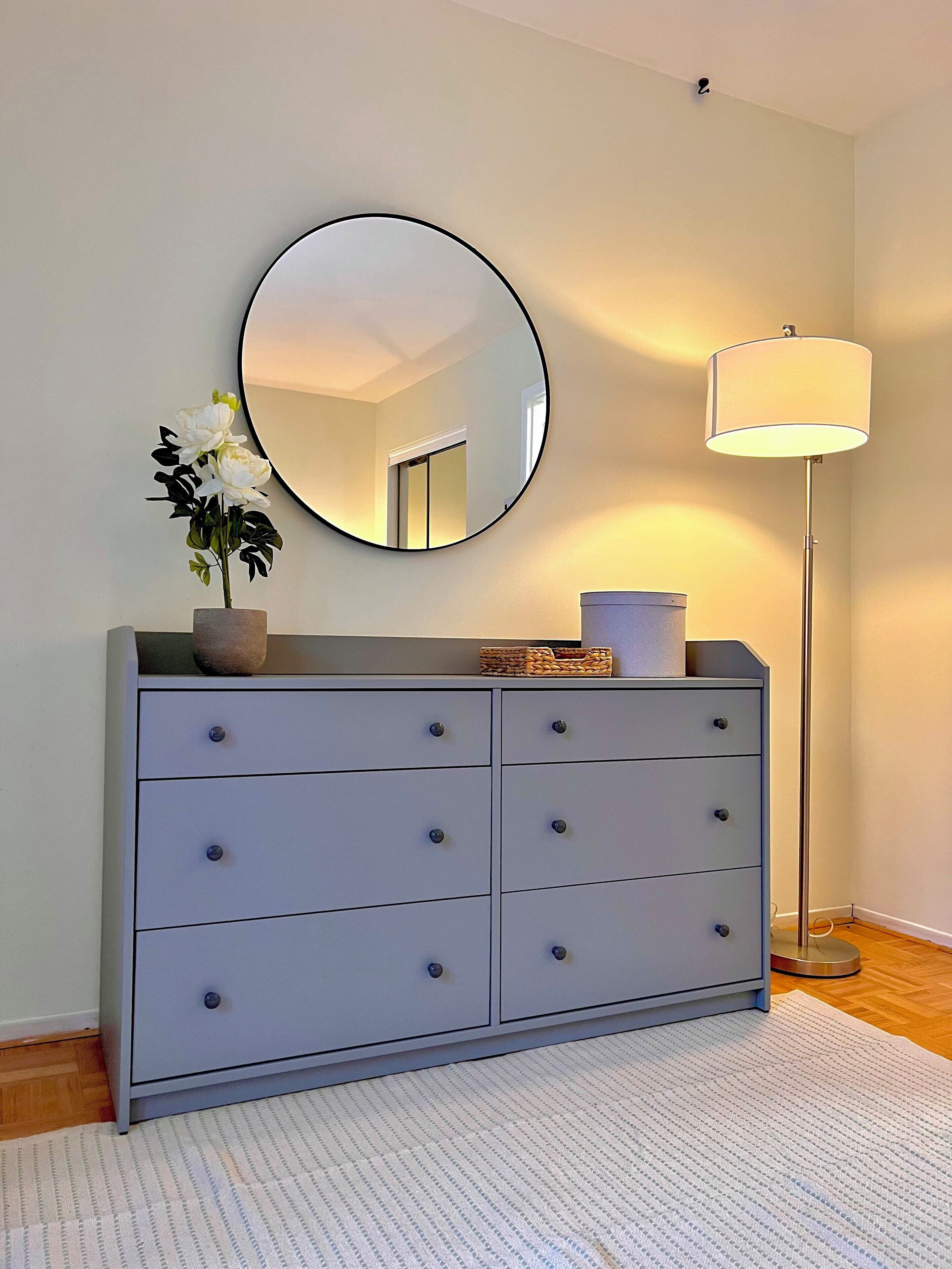 A grey dresser with a round mirror above it, a white vase with flowers, and a floor lamp to the right.