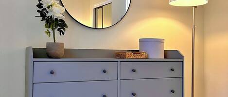 A grey dresser with a round mirror above it, a white vase with flowers, and a floor lamp to the right.