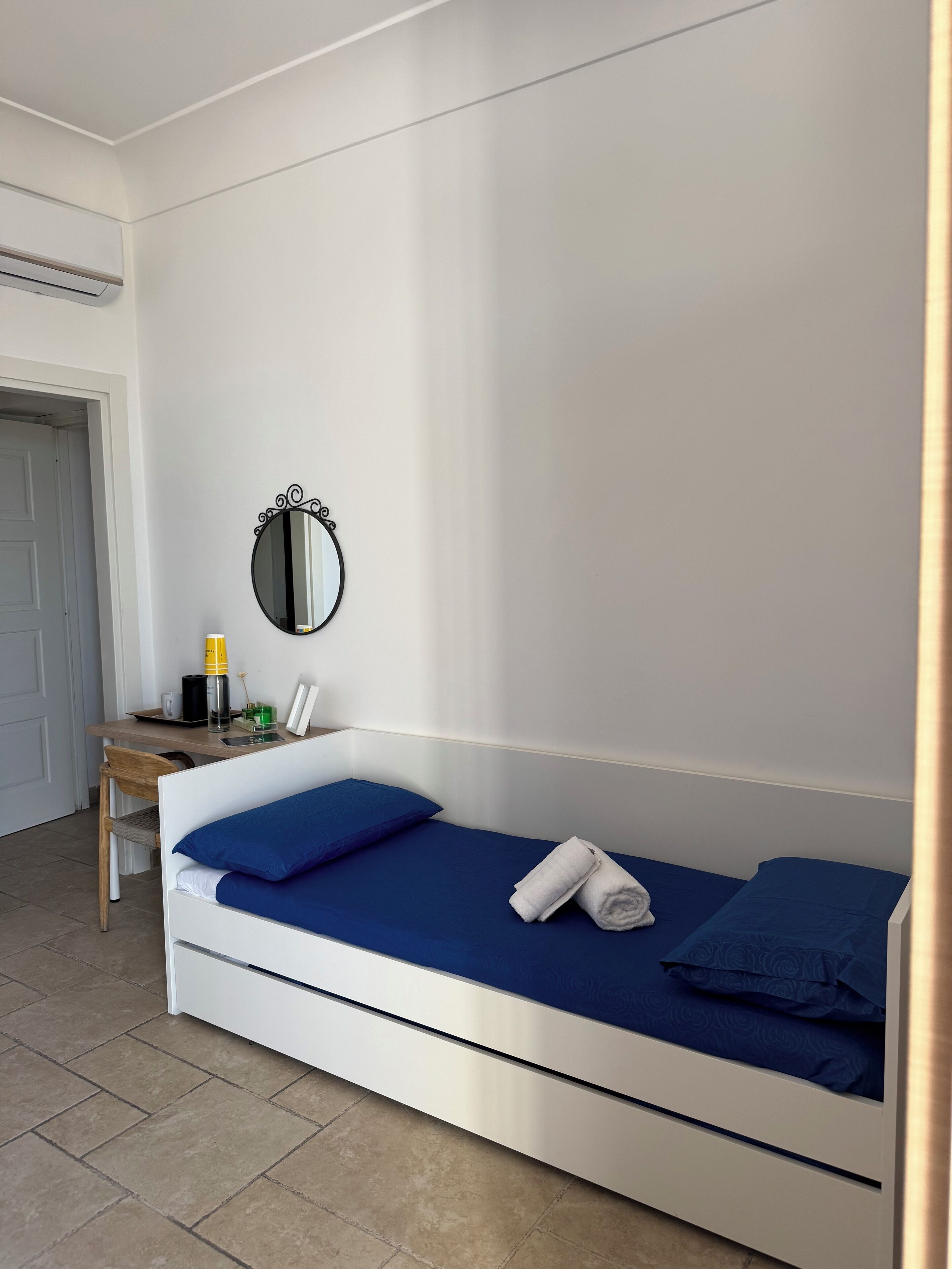 Une chambre avec un lit simple, une tête de lit blanche, une literie bleue, une petite table de chevet avec une lampe, un miroir et une porte située à gauche.