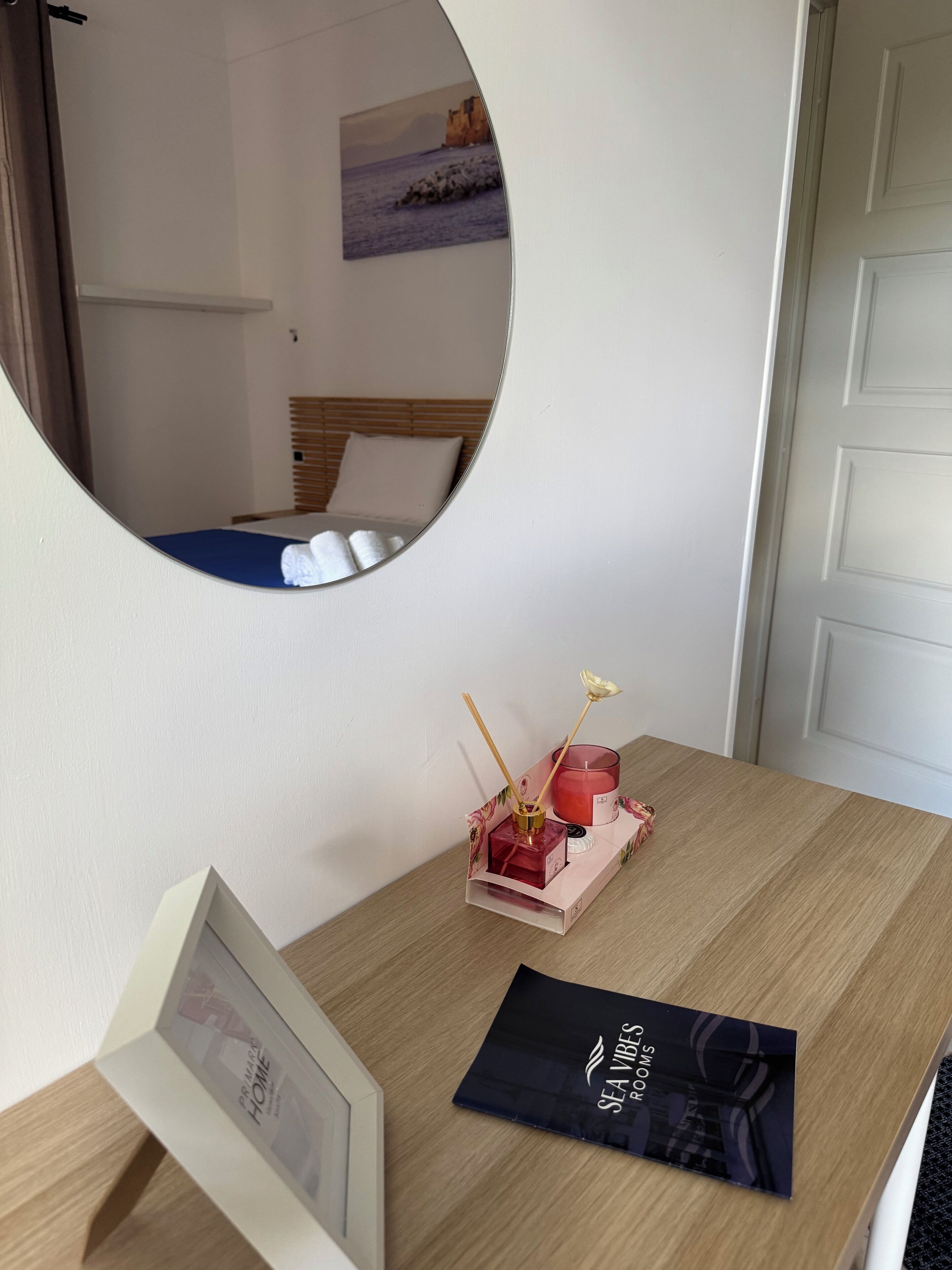 A room with a wooden desk, a mirror, a framed picture, a small table with a candle and a decorative item, and a door.