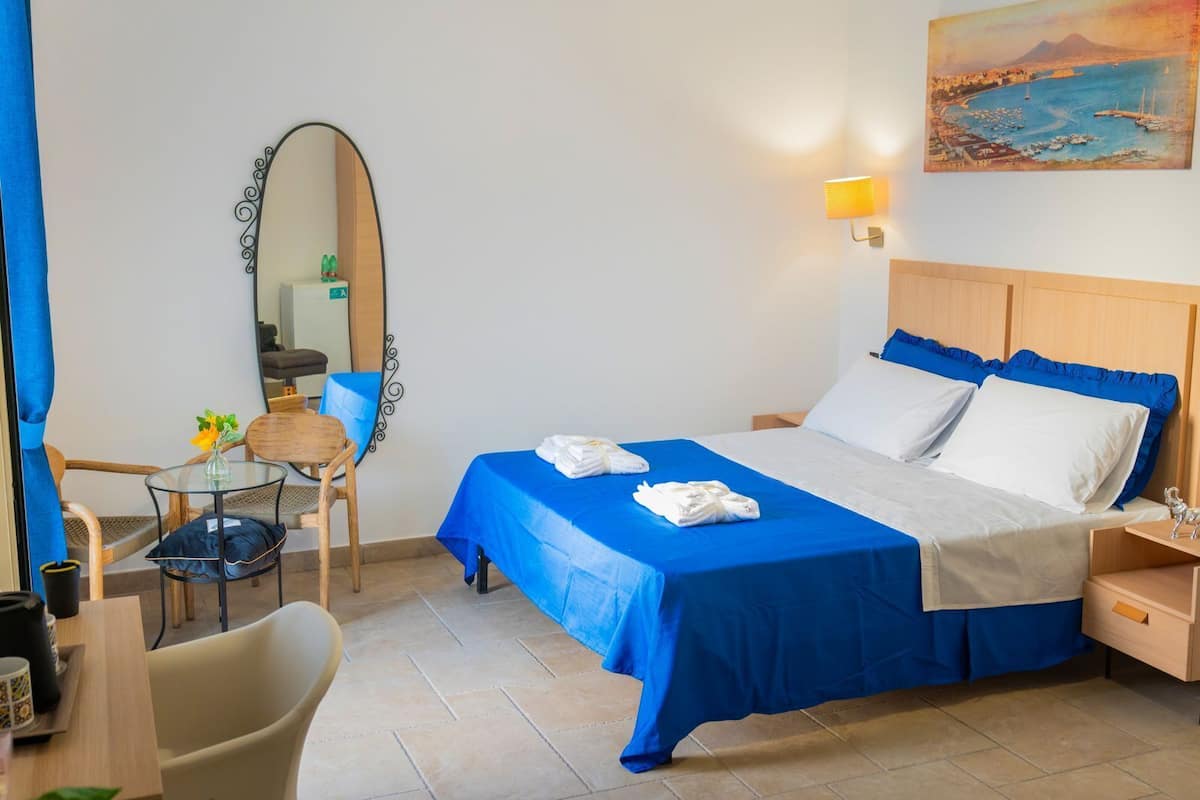 A hotel room with a bed, a desk, a chair, a mirror, and a painting on the wall.
