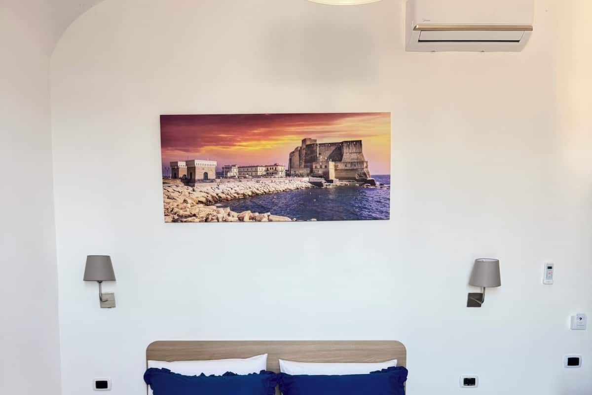 A bedroom with a large bed, two bedside tables, a wall-mounted air conditioner, and a painting of a coastal scene.