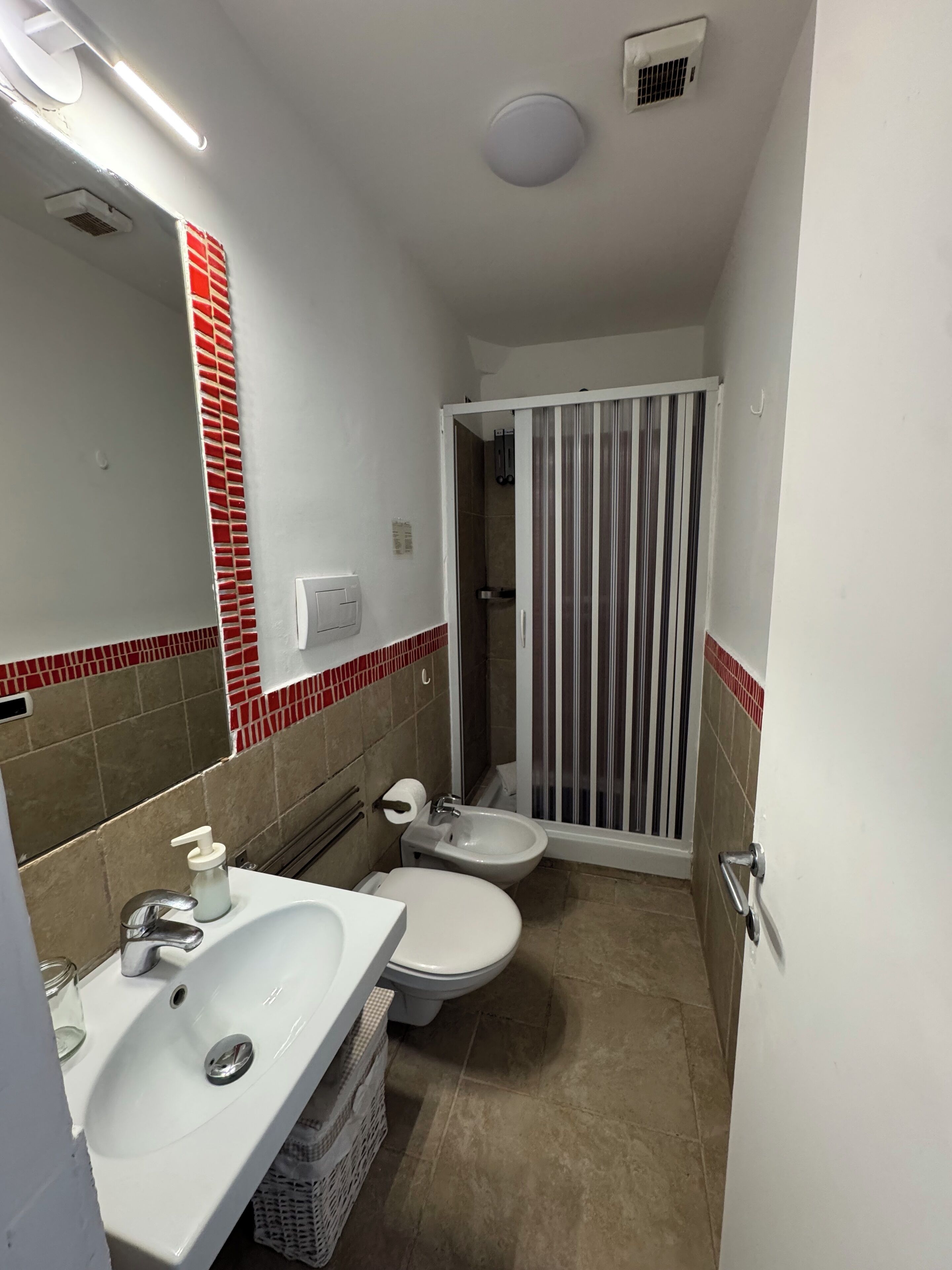 A compact bathroom with a white sink, toilet, and bidet, a shower with a striped curtain, and a red-tiled backsplash.