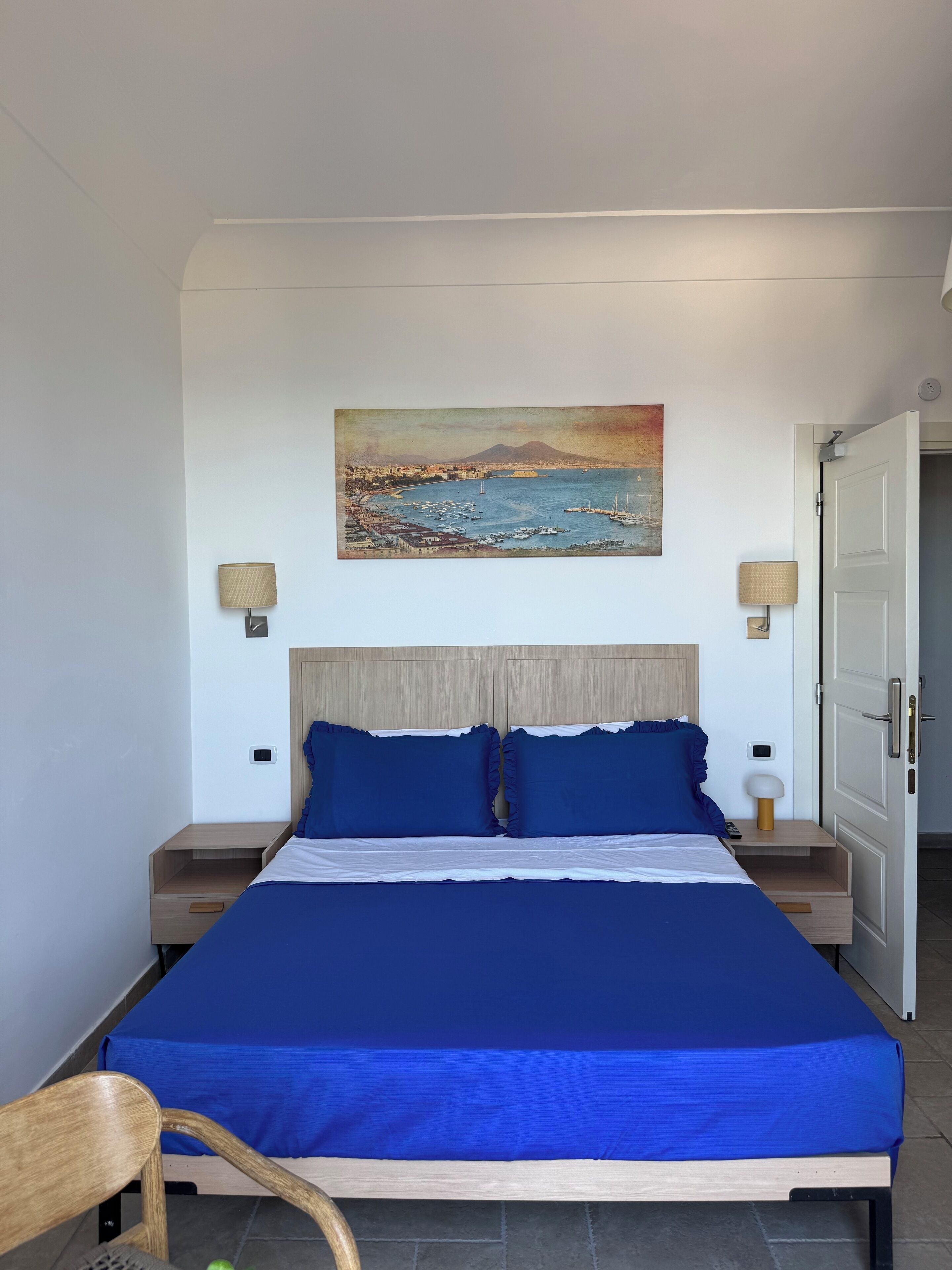 A bedroom with a bed, two bedside tables, a nightstand, a chair, a painting, and a door.
