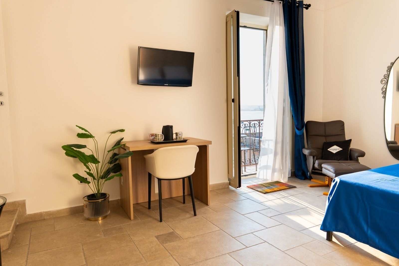 Une chambre d’hôtel équipée d’un bureau, d’une chaise, d’un lit, d’une télévision, d’un balcon et d’une plante en pot.