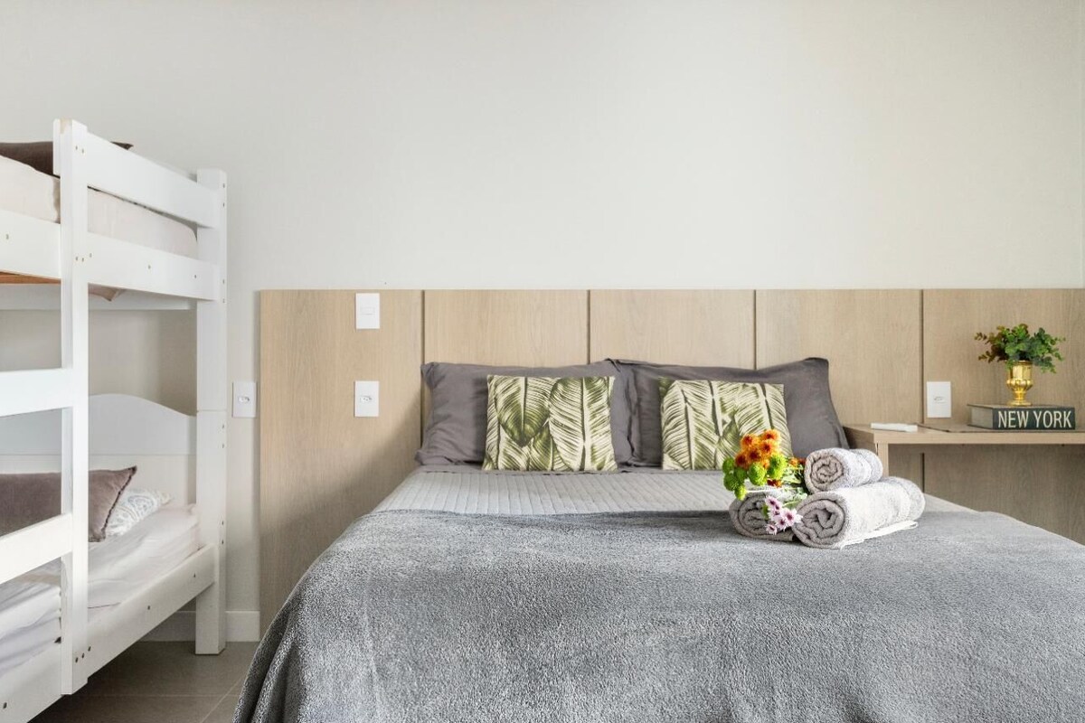 A hotel room with a bunk bed, a single bed with a grey blanket, and a bedside table with a vase of flowers and rolled towels.