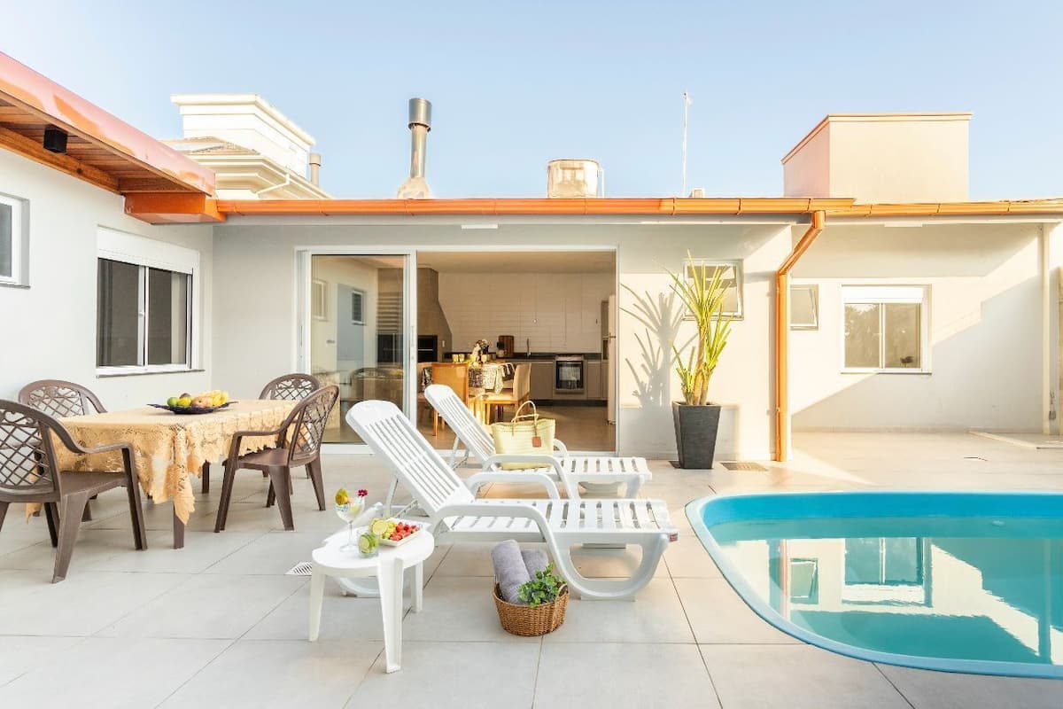 Zona junto a la piscina con sillones de descanso, una mesa de comedor y una cocina visible a través de una puerta abierta.