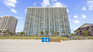 A high-rise hotel with multiple balconies, located near a beach with a sign indicating the number 14.