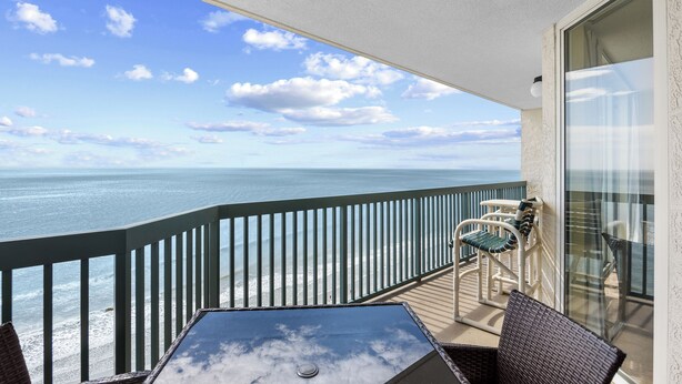 A balcony with a view of the ocean, a table, and two wicker chairs.