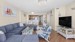 A living room with a sofa, chairs, a coffee table, a TV, and a kitchen in the background.