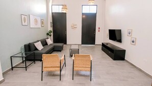 A modern living room with a sofa, two wicker chairs, a glass coffee table, a TV, and artwork on the walls.