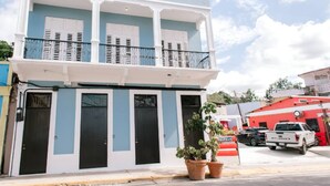 A two-story building with a balcony, a red vehicle parked in front, and potted plants on the sidewalk.