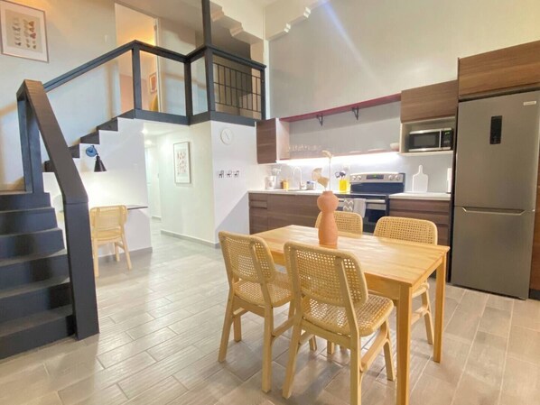A modern kitchen with a dining area, featuring a staircase, wooden chairs, and a stainless steel refrigerator.