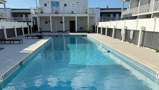 A rectangular swimming pool with a tiled edge, surrounded by a white building with balconies and a covered walkway. - Charming Apartment in Gorgeous George Town with GYM, Pool, and Pickleball Court (George Town)
