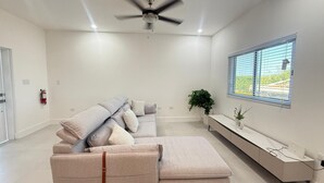 A modern living room with a grey sectional sofa, a ceiling fan, a window with blinds, a small plant, and a white console table. - Charming Apartment in Gorgeous George Town with GYM, Pool, and Pickleball Court (George Town)