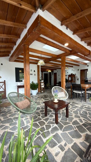 Un'ampia area interna con pavimento in pietra, travi in legno a soffitto e un angolo salotto con una poltrona in vimini e un tavolo.