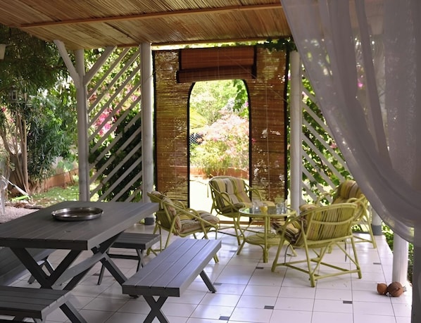 An outdoor patio with picnic tables and wicker chairs, surrounded by a bamboo fence and greenery.