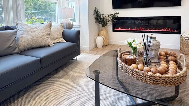 Ein modernes Wohnzimmer mit großem Flachbildfernseher über einem Kamin, einer dunkelgrauen Couch mit Kissen und einem Glastisch mit Dekoration.
