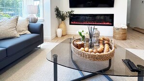 Ein modernes Wohnzimmer mit einem großen Flachbildfernseher an der Wand, einem dunklen Sofa, einem Glastisch mit Dekoration und einem Kamin unter dem Fernseher.