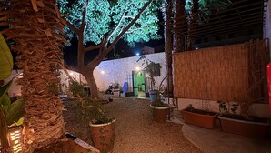 Un espacio exterior nocturno con un sendero de grava, plantas en macetas y un edificio con pared de bambú al fondo.