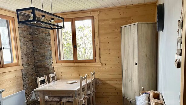 A cozy cabin interior with a wooden dining table, chairs, a stone wall, and a loft bed with pillows.