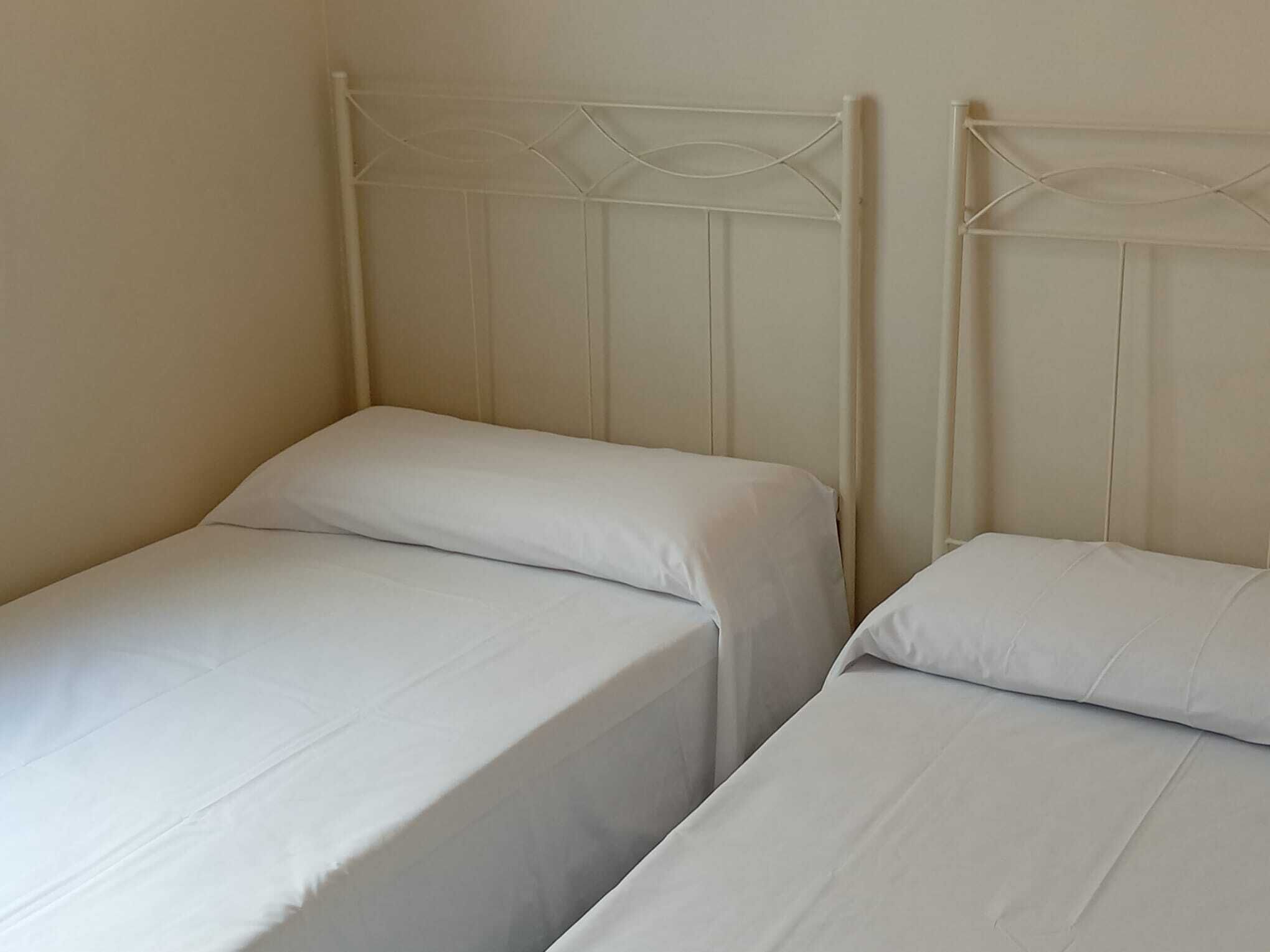 Two single beds with white linens in a room with plain walls and a simple metal headboard design.