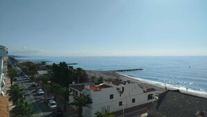 Ein Küstenblick mit blauem Himmel, einem Strand und einer Straße gesäumt von Gebäuden und parkenden Autos.