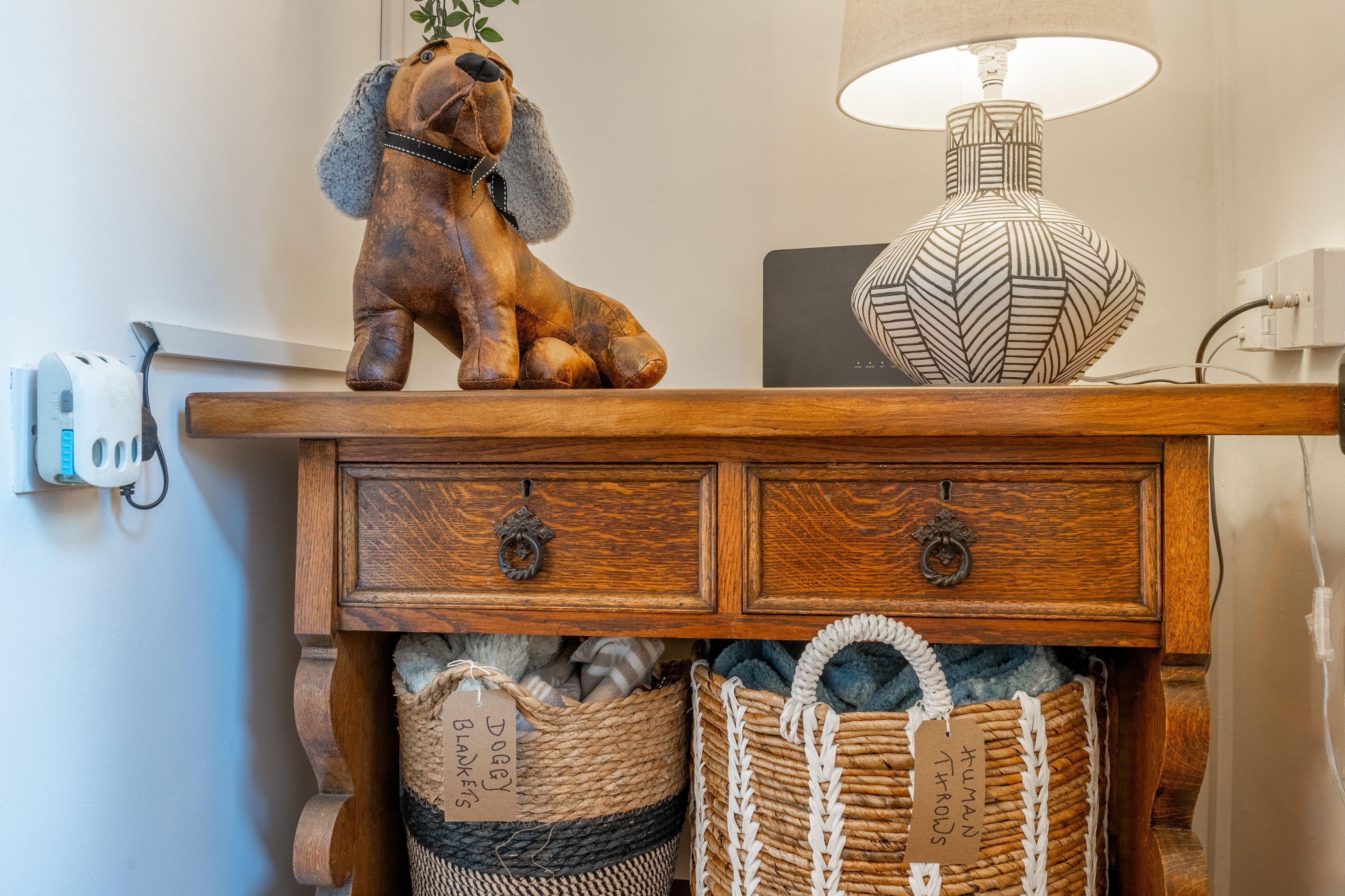 A wooden console table with two drawers, a decorative dog figurine, and two baskets labeled 'DOGGY BLANKETS' and 'HUMAN THROW.'