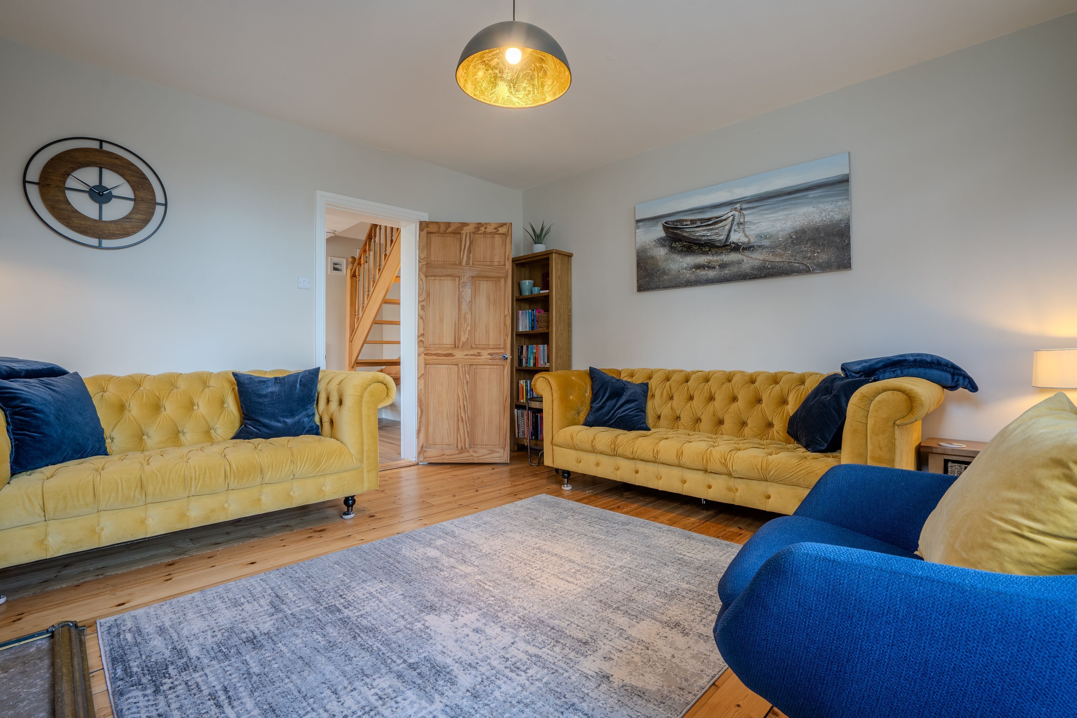 Un salon avec deux canapés jaunes, un fauteuil bleu, un escalier en bois et une bibliothèque.