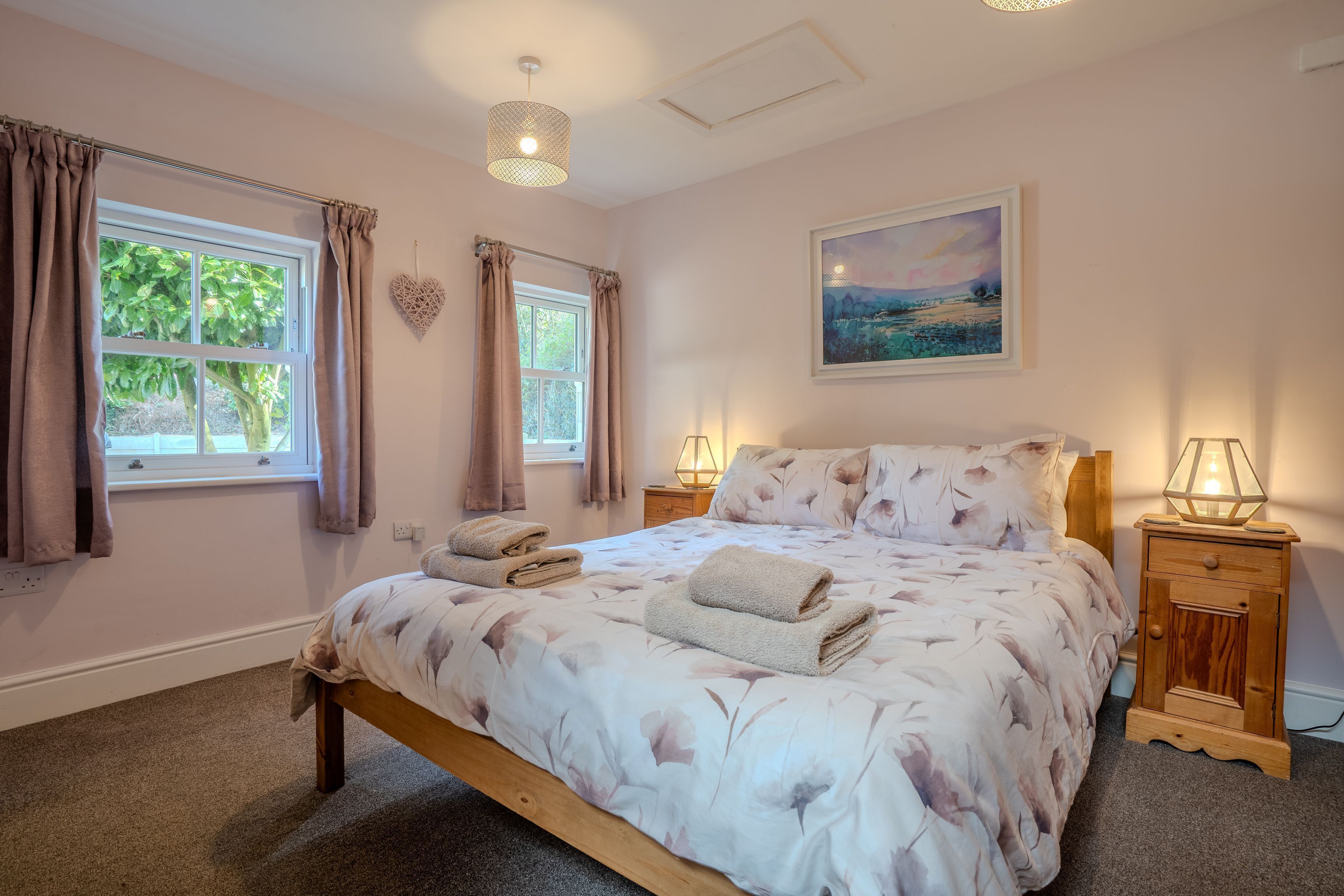 Une chambre à coucher avec un lit, deux tables de chevet, une fenêtre avec des rideaux, un tableau au mur et une lampe sur la table de chevet.