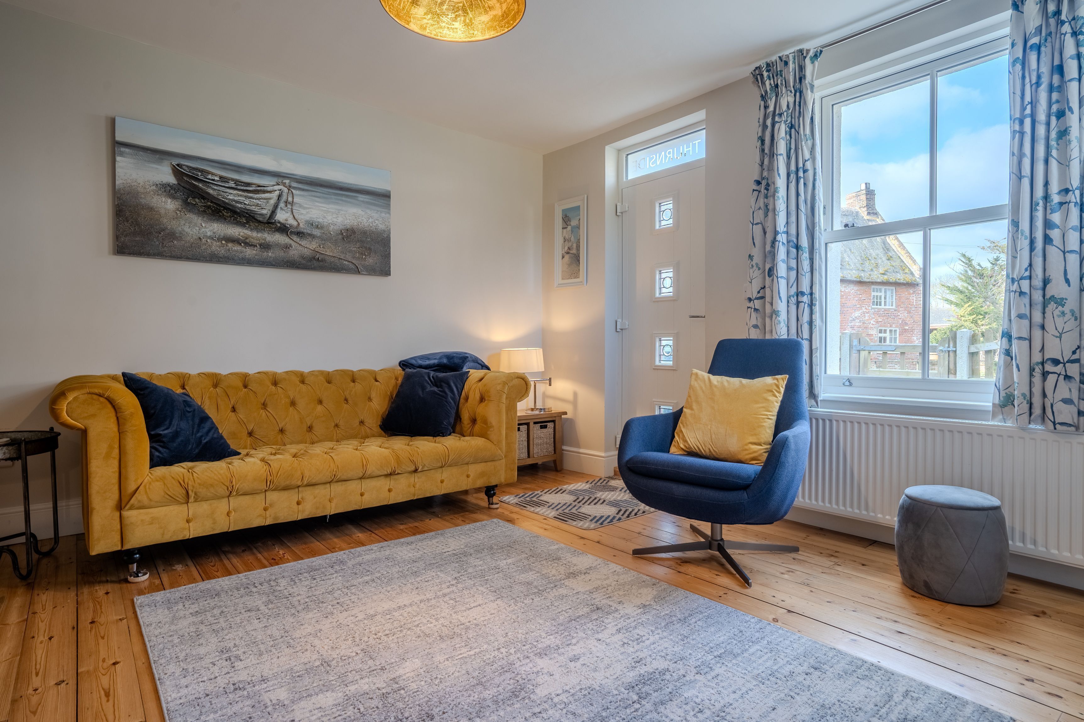 Un salon avec un canapé jaune, un fauteuil bleu, un tableau représentant un bateau et une fenêtre avec des rideaux.