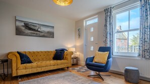 Una sala de estar con un sofá amarillo, un sillón azul, un cuadro de un barco y una ventana con cortinas.