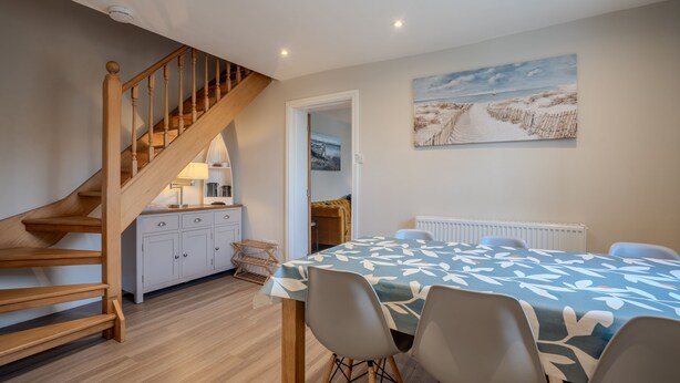Un interior moderno con una escalera de madera, una zona de comedor con una mesa y sillas, una cocina con armarios y un cuadro en la pared.