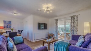 Uma sala de estar moderna com um sofá roxo, televisão de tela plana, área de jantar com mesa e cadeiras, e vista para o exterior por uma porta de vidro.