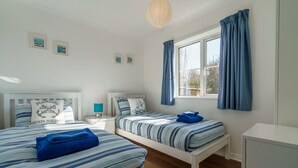 Quarto com duas camas, janela com cortinas, mesa de cabeceira com abajur e parede com quadros emoldurados.