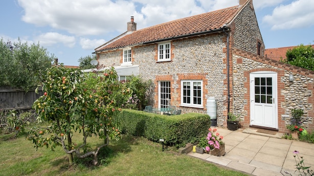 A stone house with a tiled roof, a front door, and a small garden with a tree and flowers. - Sunny Cottage by East Ruston Cottages (Southrepps)