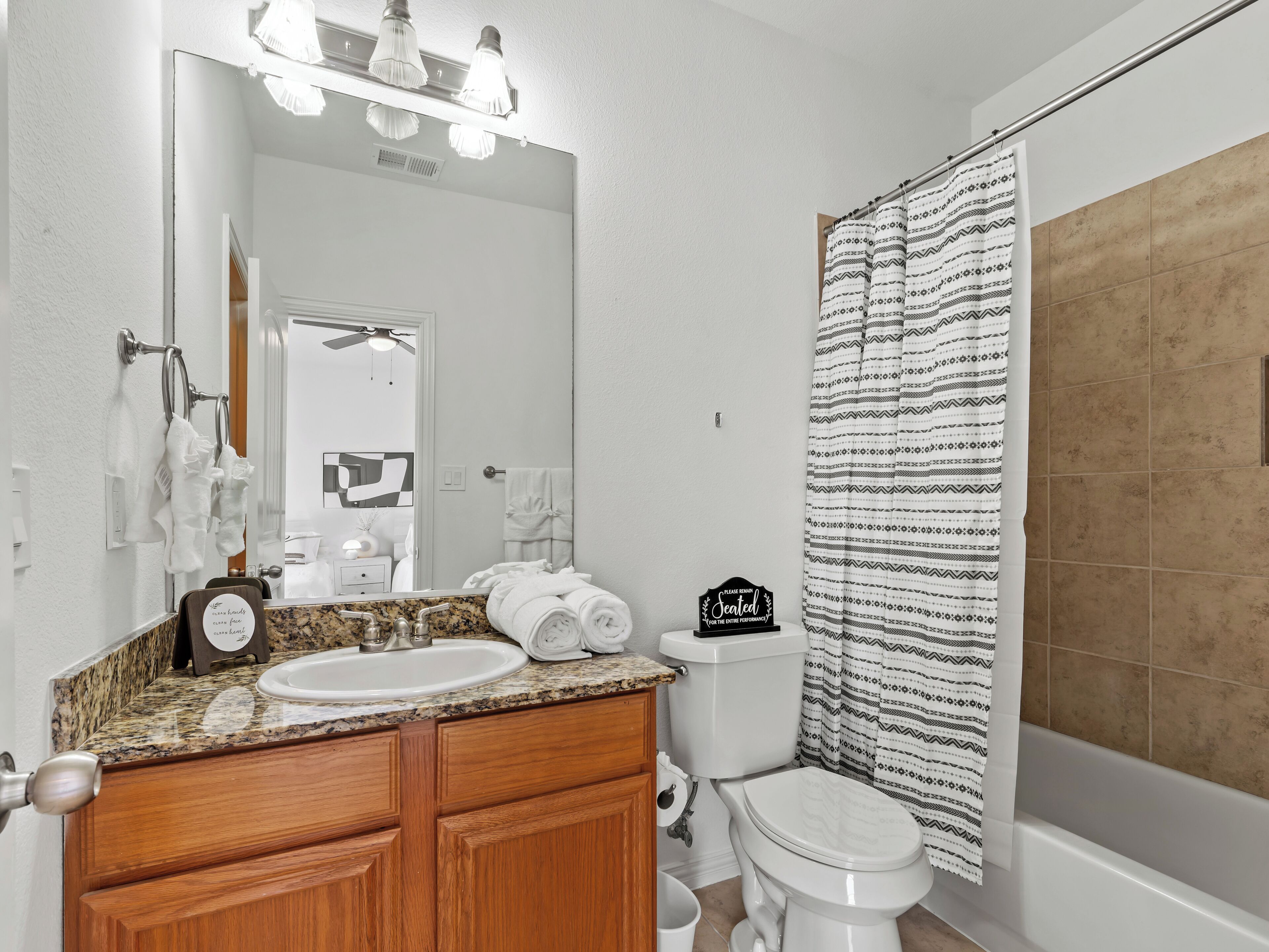 Une salle de bain avec un lavabo sur un plan de travail en granit, des meubles en bois, des toilettes blanches et une douche avec des rideaux à motifs.