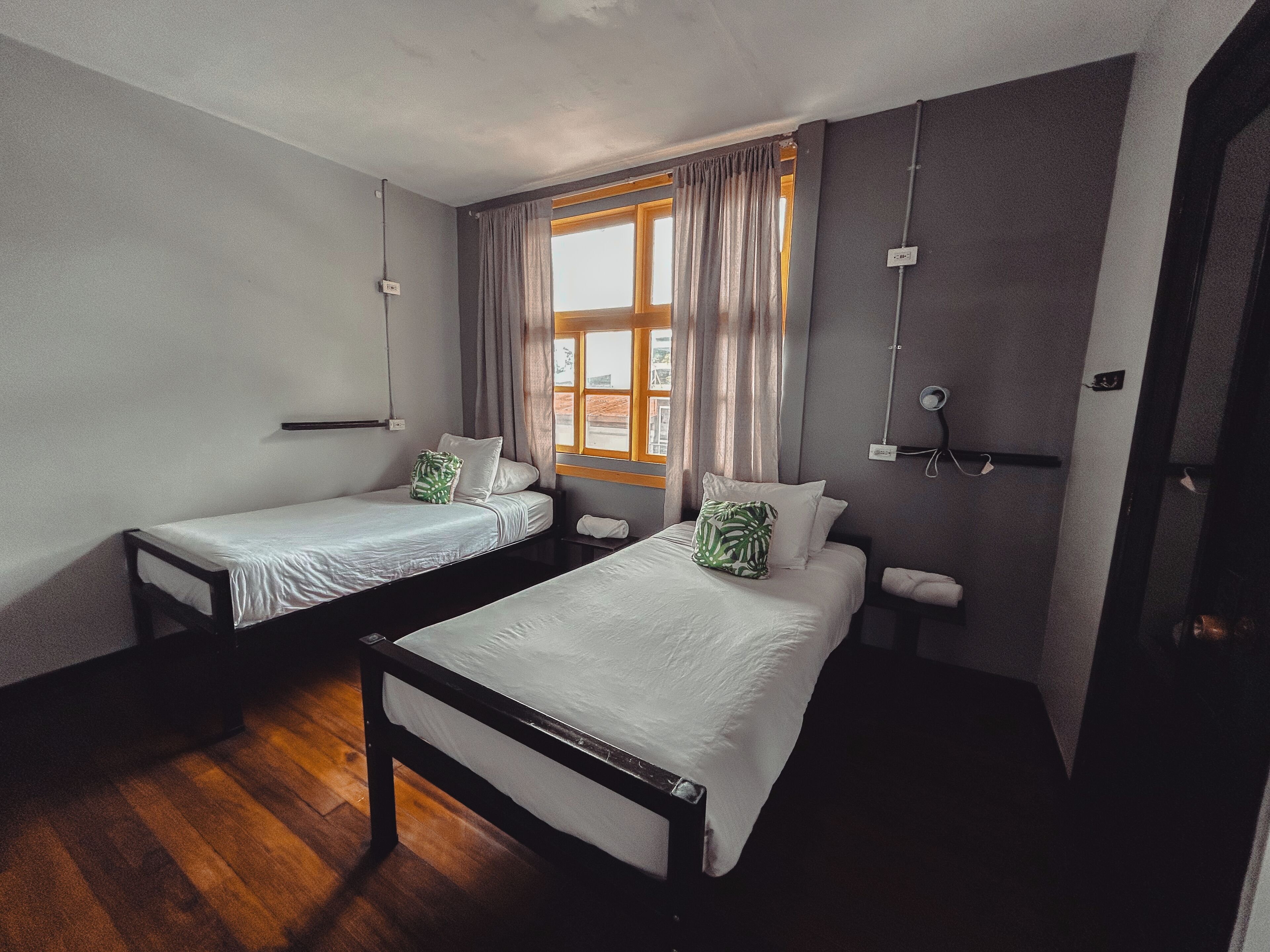 A room with two single beds, a window with curtains, a wooden floor, and a wall-mounted light fixture.