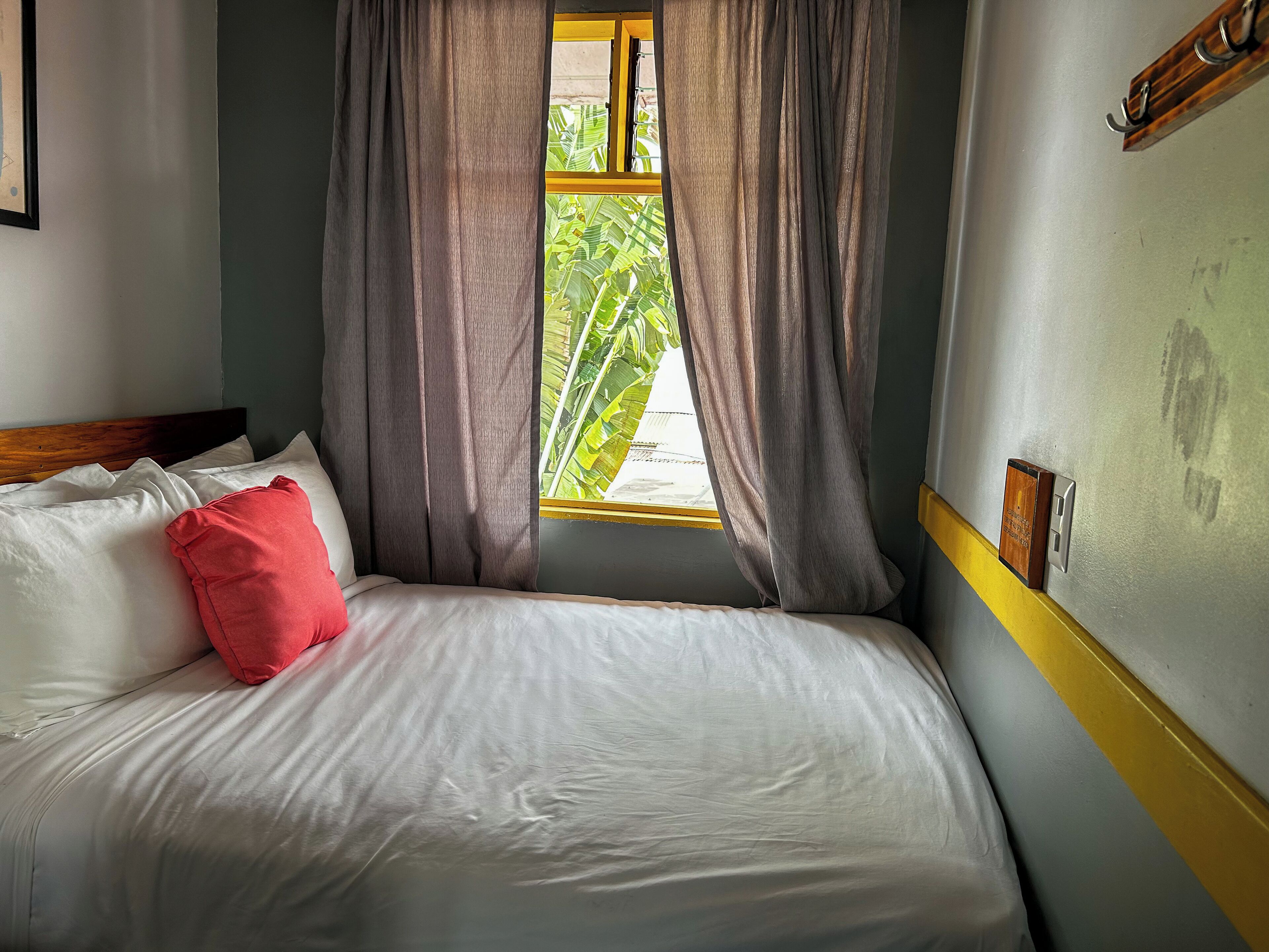 Una cama con ropa de cama blanca y una almohada roja, una ventana con cortinas y una pared con interruptor de luz y un panel de madera.