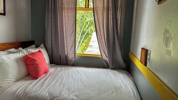 Una cama con ropa de cama blanca y una almohada roja, una ventana con cortinas y una pared con interruptor de luz y un panel de madera.