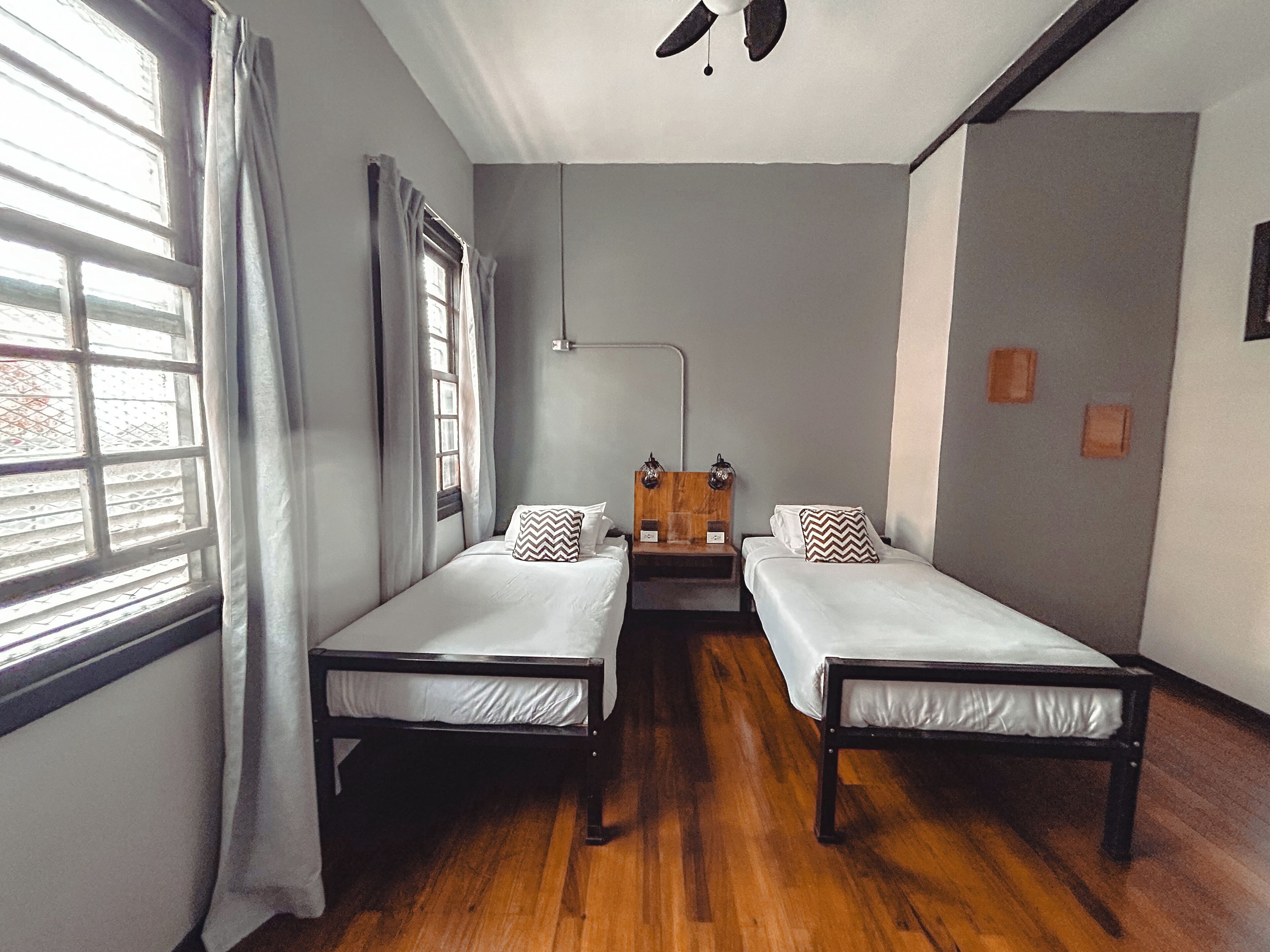 A room with two single beds, a wooden headboard, a bedside table with a lamp, and a window with curtains.