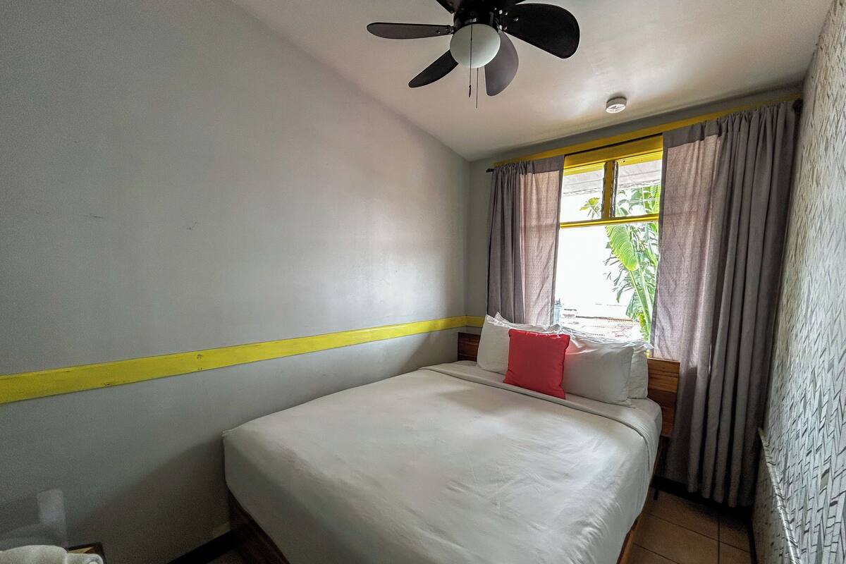 Una habitación de hotel con cama, ventilador de techo, ventana con cortinas y una franja amarilla en la pared.
