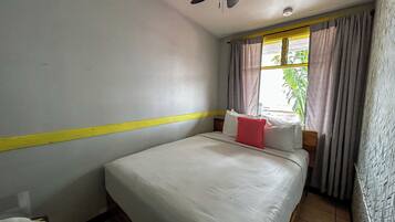 Una habitación de hotel con cama, ventilador de techo, ventana con cortinas y una franja amarilla en la pared.