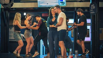 A group of people dancing in a room with a large screen displaying a video, a sign with 'COV' partially visible, and a circular sign with a camera icon and text.