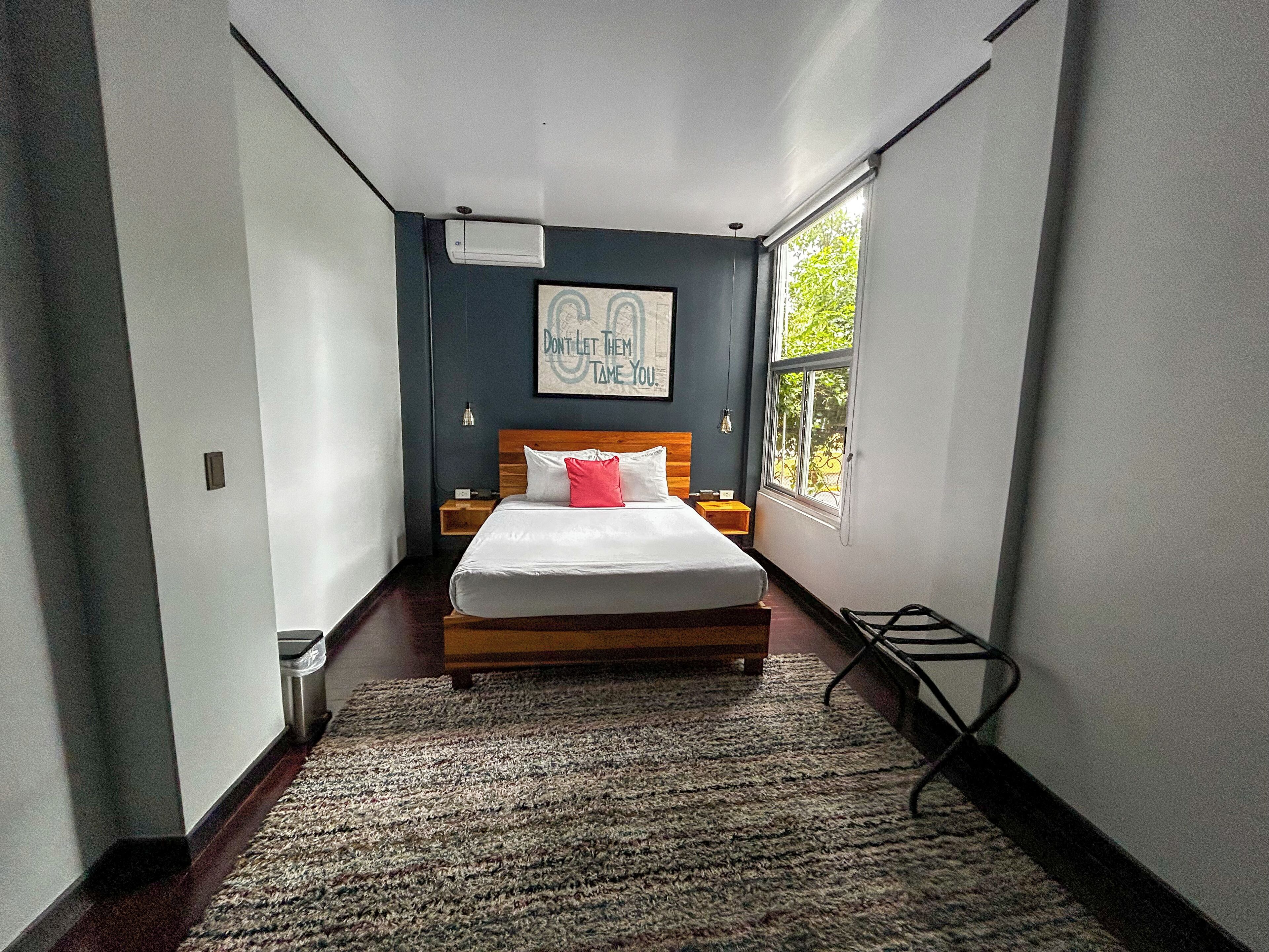 Habitación de hotel estrecha con una cama de madera, una mesita de noche pequeña, un buró, aire acondicionado montado en la pared, una cita inspiradora enmarcada y una ventana con vistas a vegetación.
