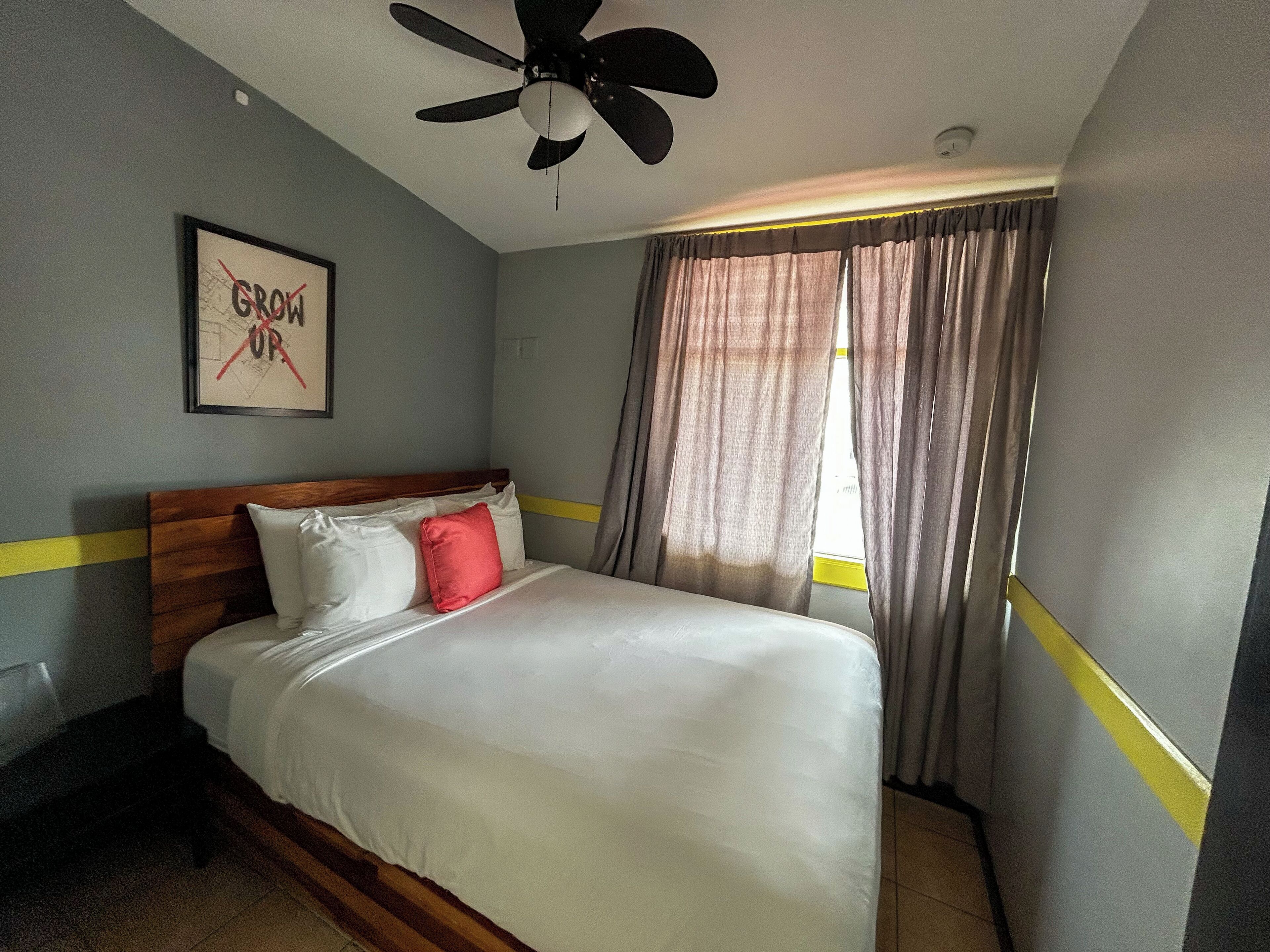 Une chambre d’hôtel avec un lit, un ventilateur de plafond, une fenêtre avec des rideaux et un mur orné d’un tableau.