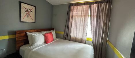 Une chambre d’hôtel avec un lit, un ventilateur de plafond, une fenêtre avec des rideaux et un mur orné d’un tableau.