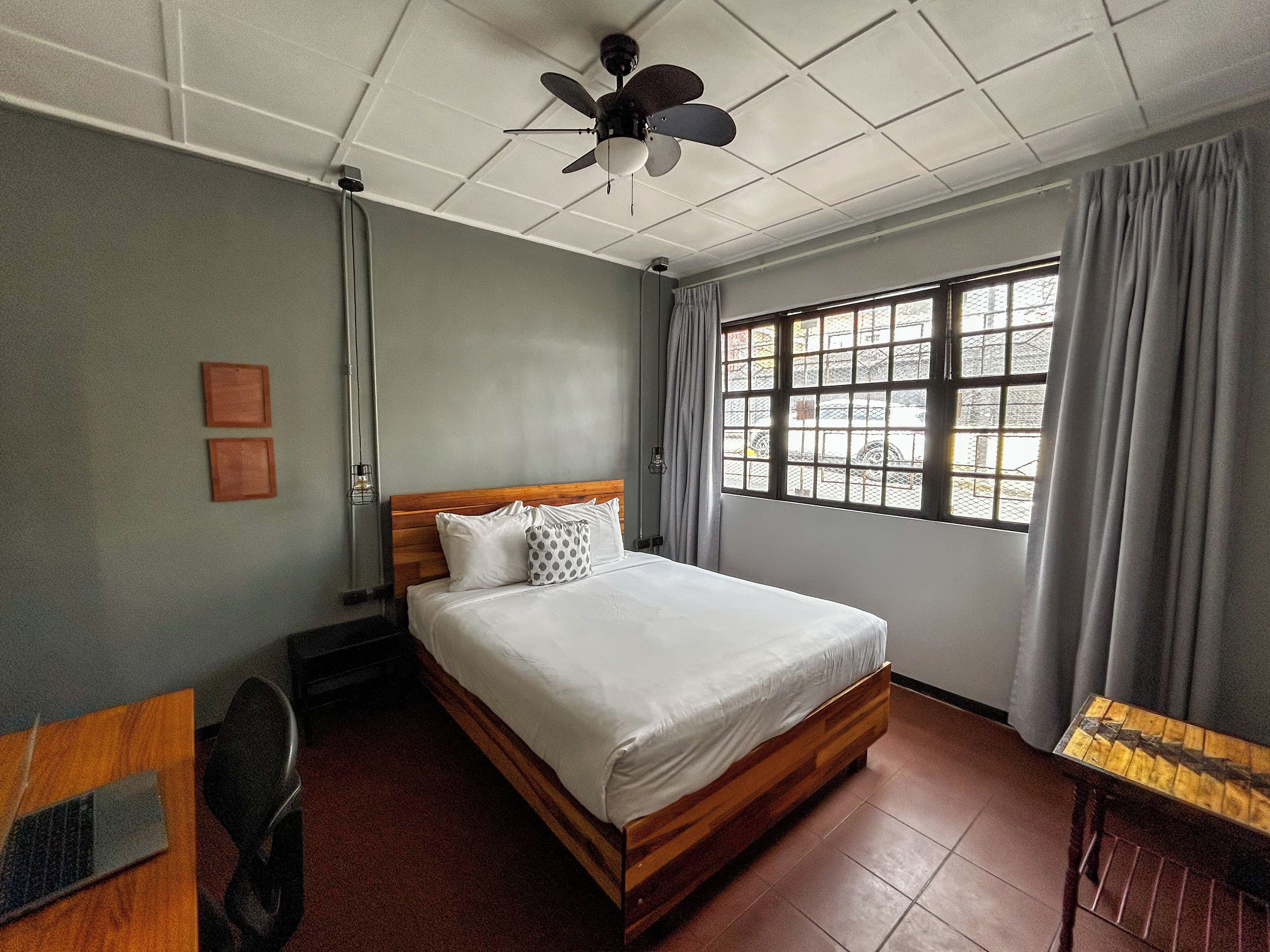 A hotel room with a bed, a desk, a chair, a ceiling fan, a window with curtains, and a view of the outside.
